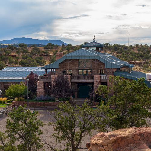 This image shows a large, rustic building set in a desert landscape with trees, mountains in the background, and a cloudy sky overhead.