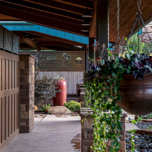 A covered walkway with hanging plants and wooden walls leads to a landscaped garden area with rocks and greenery.
