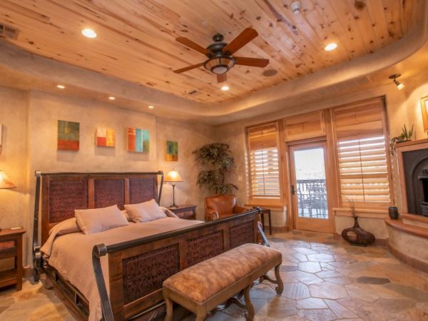 A cozy bedroom with a wooden ceiling, fan, bed, bench, lamps, abstract art, and fireplace, featuring a stone floor and window doors.