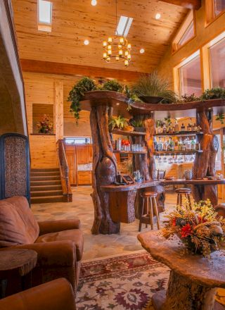 A cozy, rustic living room with wooden furniture, plants, and a bar area, featuring warm lighting and a staircase leading upwards.