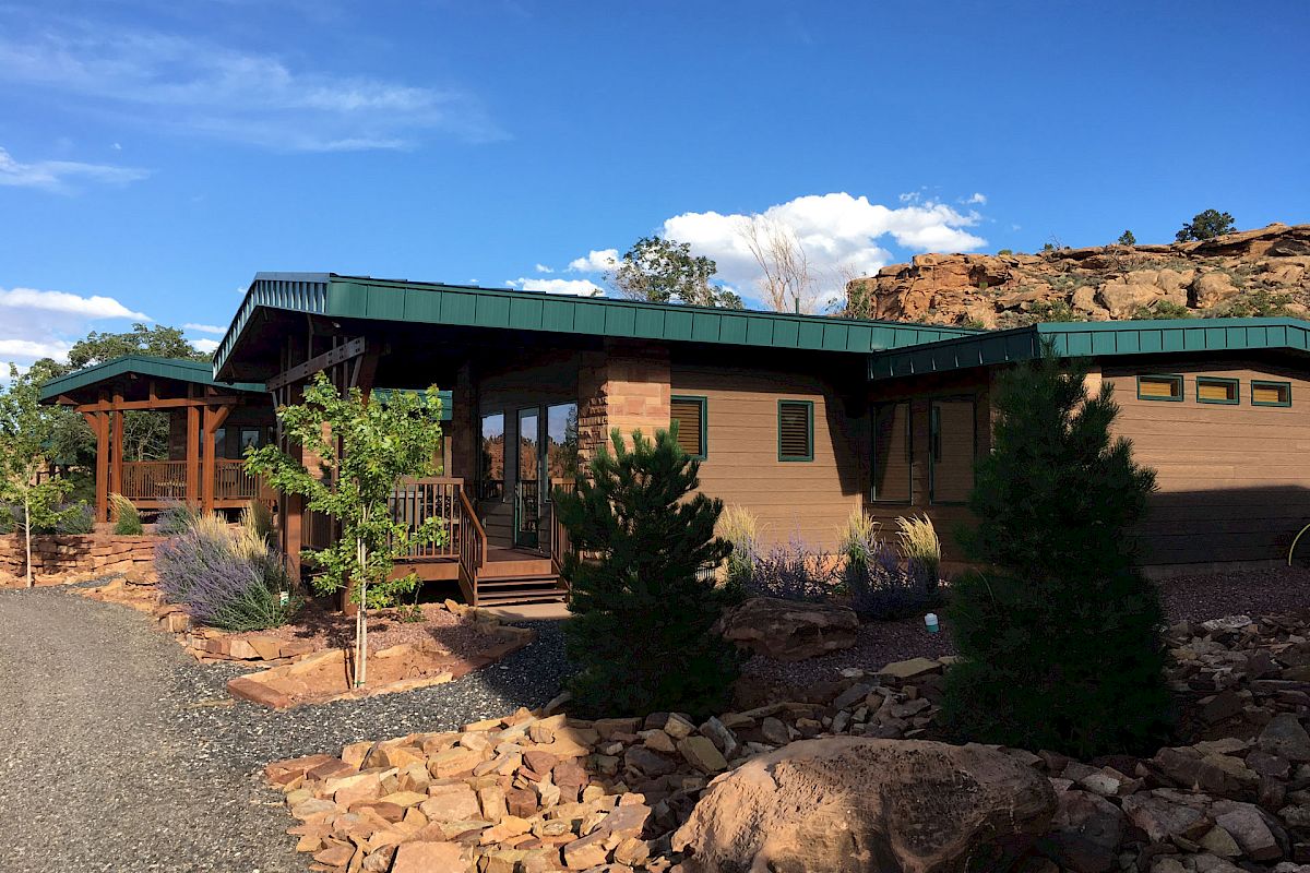 A modern, single-story building with a green roof, surrounded by rocks and trees under a clear blue sky.