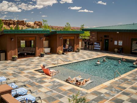A sunny outdoor pool area with people relaxing by the water, surrounded by buildings with a desert landscape in the background.