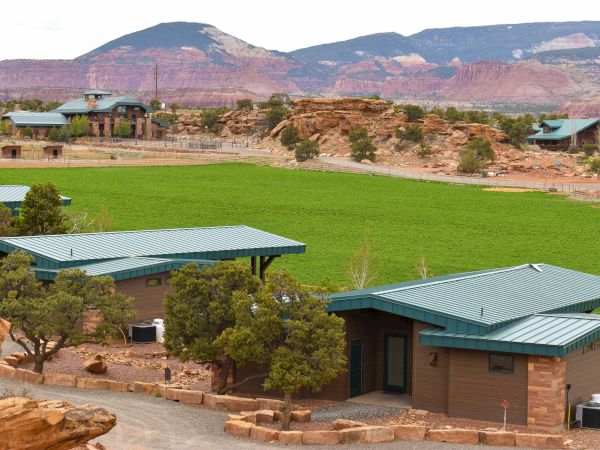 The image shows cabins with green roofs near a grassy area, surrounded by trees and mountains under a partly cloudy sky.