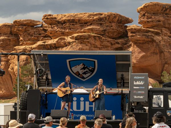The image shows two musicians performing on a small outdoor stage with rocky formations in the background, in front of a gathered crowd.