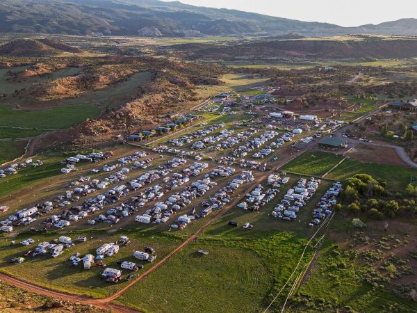 An aerial view of a large campsite with numerous vehicles and tents, set in a hilly, open landscape under a clear sky.