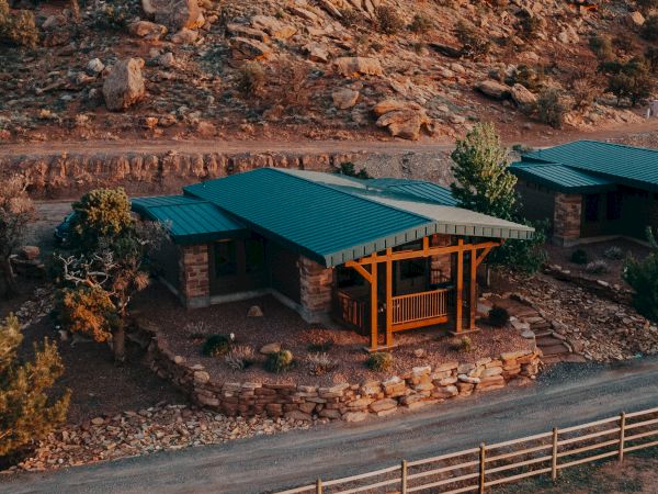 A rugged canyon landscape with a lodge-like building featuring green metal roofs, overlooking rocky cliffs and distant hills at dusk.