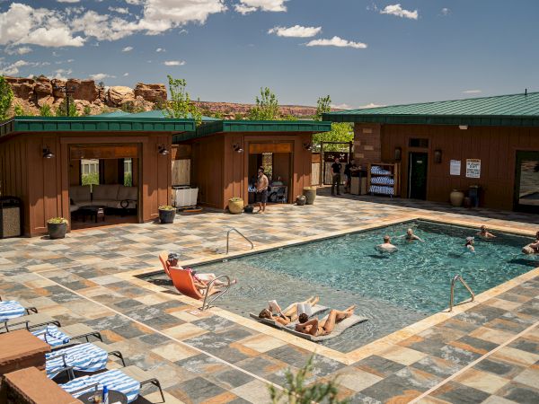 A sunny hotel pool area with a rectangular pool, people lounging on chairs, and low-rise brick buildings with green roofs in a desert setting.