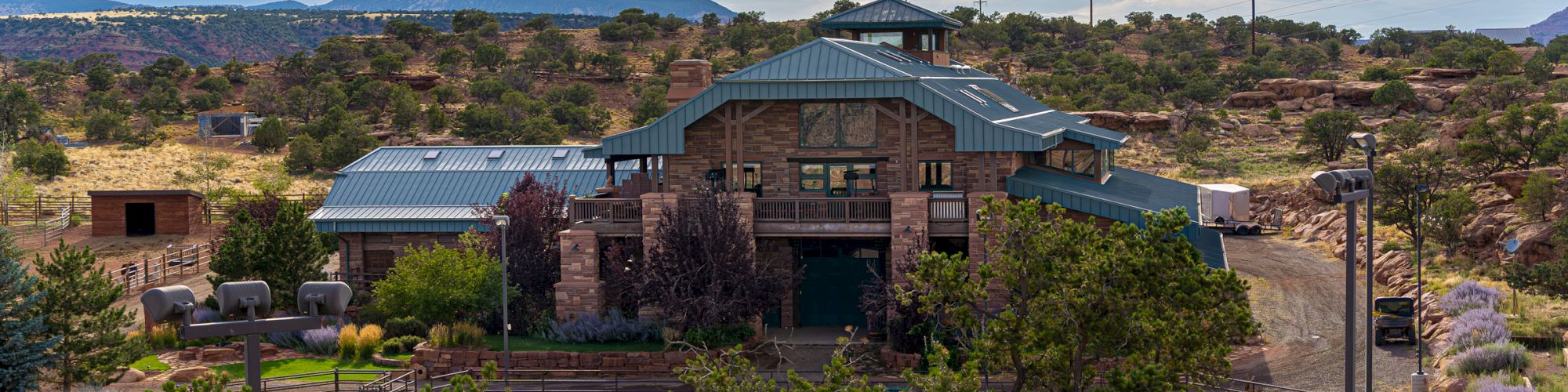 A modern lodge-style building sits among desert shrubs and rocks, with a wide entrance, metal roof, and distant mountains under a cloudy sky.