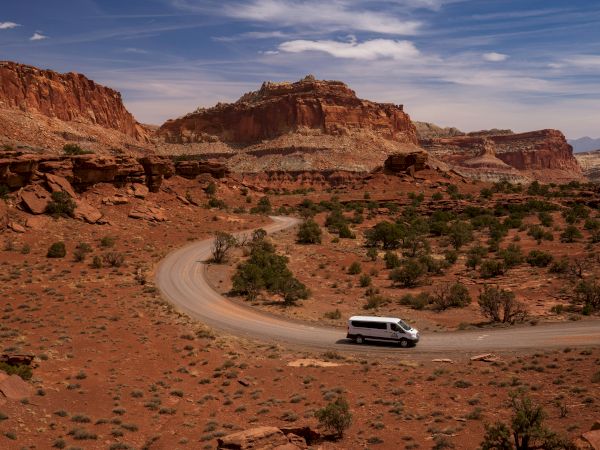 A white van drives along a winding road through a red desert canyon with rocky cliffs and sparse green shrubs, under a blue sky.