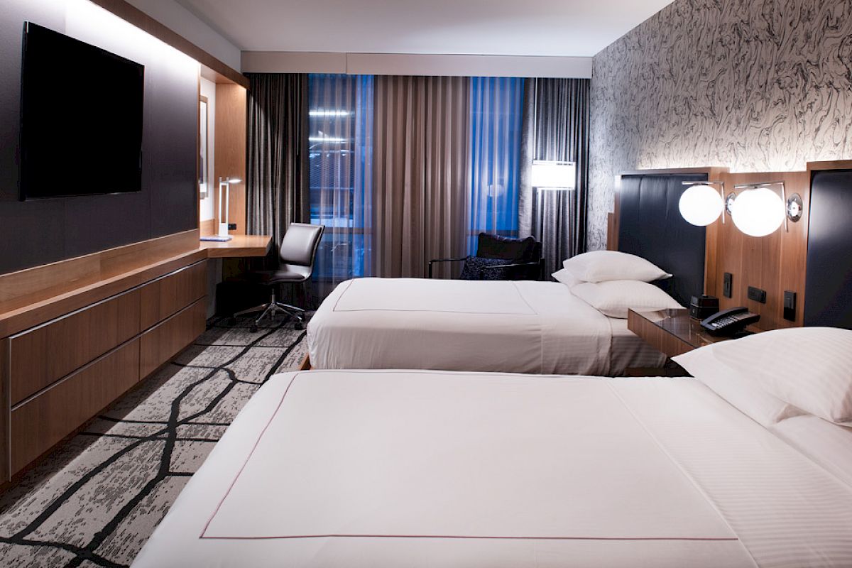 A modern hotel room with two beds, a wall-mounted TV, work desk, and ambient lighting. The decor features neutral tones and patterned carpet.