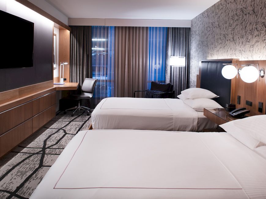 A modern hotel room with two beds, a wall-mounted TV, work desk, and ambient lighting. The decor features neutral tones and patterned carpet.