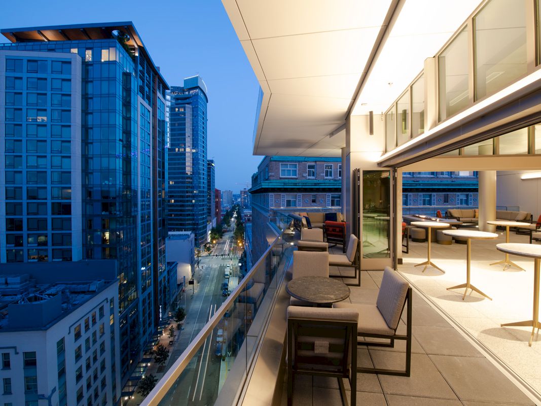 A rooftop terrace overlooking a cityscape at dusk, with seating areas and glass railings, adjacent to tall modern buildings.