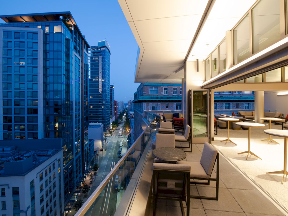 A modern rooftop terrace with seating overlooks a city skyline at dusk, featuring tall buildings and a street view.