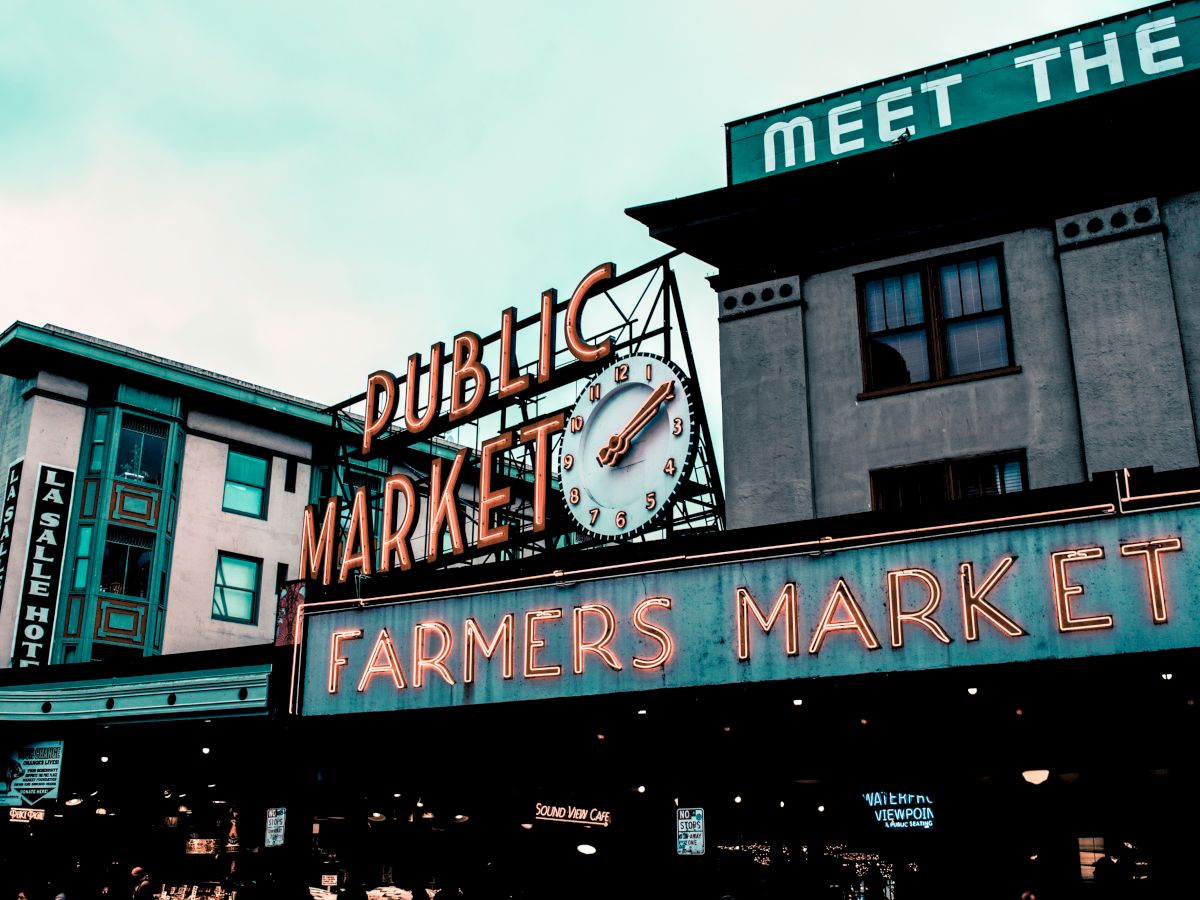 The image shows the iconic neon sign of a public farmers market with an adjacent clock, set against urban buildings.