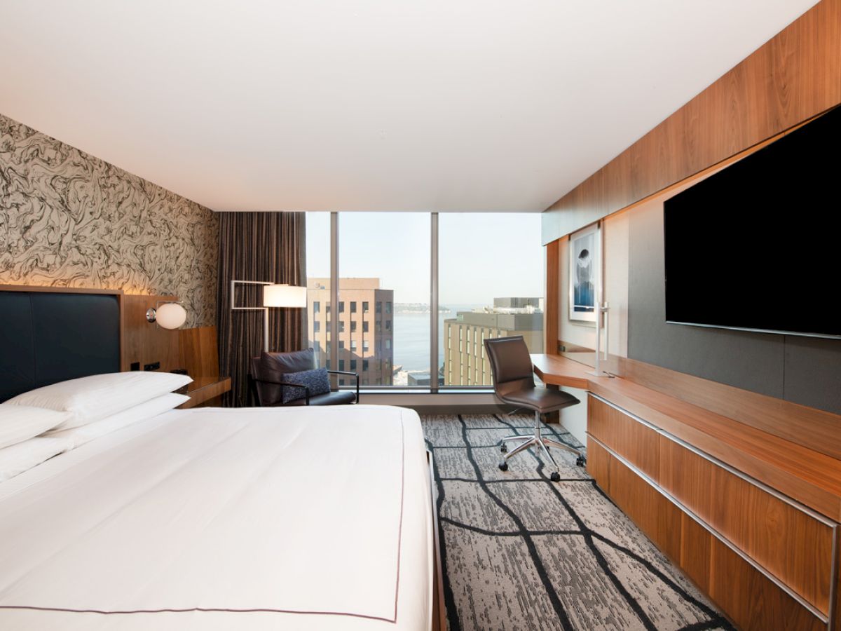 A modern hotel room with a large bed, desk, and wall-mounted TV, featuring a city view through a floor-to-ceiling window.