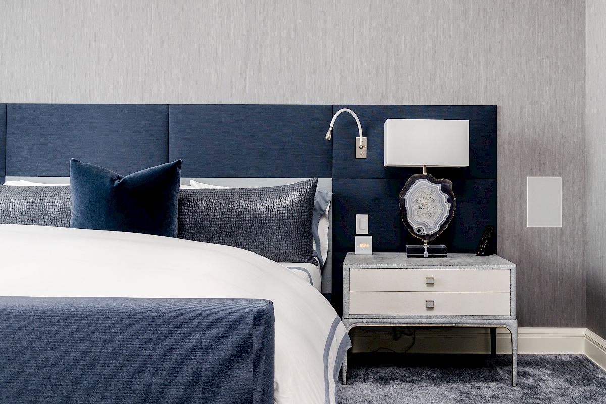 A modern bedroom with a blue and white color scheme, featuring a bed, pillows, a nightstand with a lamp, and framed artwork.