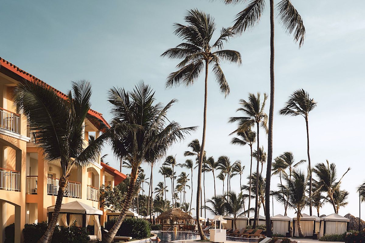 A resort pool area with palm trees, lounge chairs, and adjacent buildings under a clear sky.