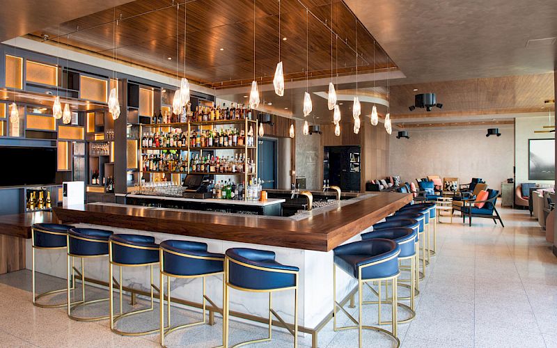 The image shows a stylish bar with a wooden counter, blue stools, pendant lighting, and a variety of bottles on shelves in a modern setting.
