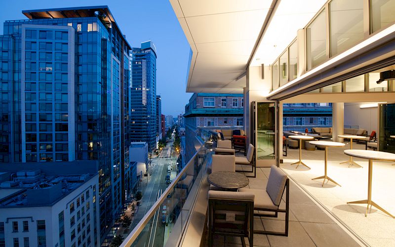 Rooftop terrace at dusk with seating, overlooking a city street and high-rise buildings under a blue evening sky.
